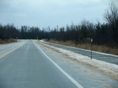 [View from the passenger seat of a car of the road and the paved bicycle trail at the edge of the road. A dusting of snow covers the grassy area.]